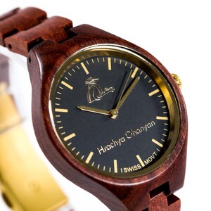 Könnte beinhalten: Eine Holzarmbanduhr mit dunklem Zifferblatt und goldfarbenen Zeigern und Akzenten. Die Uhr hat ein braunes Holzarmband und ein schwarzes Zifferblatt mit dem Text "Hrachya Chanyan" und "SWISS MOVT".