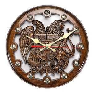 May include: A round wooden clock with a detailed carving of a coat of arms featuring an eagle and a lion. The clock has a dark brown frame with silver accents and gold clock hands. The clock face is white.