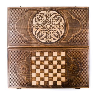 May include: A wooden backgammon and chess set. The dark brown wooden case is intricately carved with geometric and floral designs. The backgammon board is open, revealing a checkerboard pattern and a detailed central design.