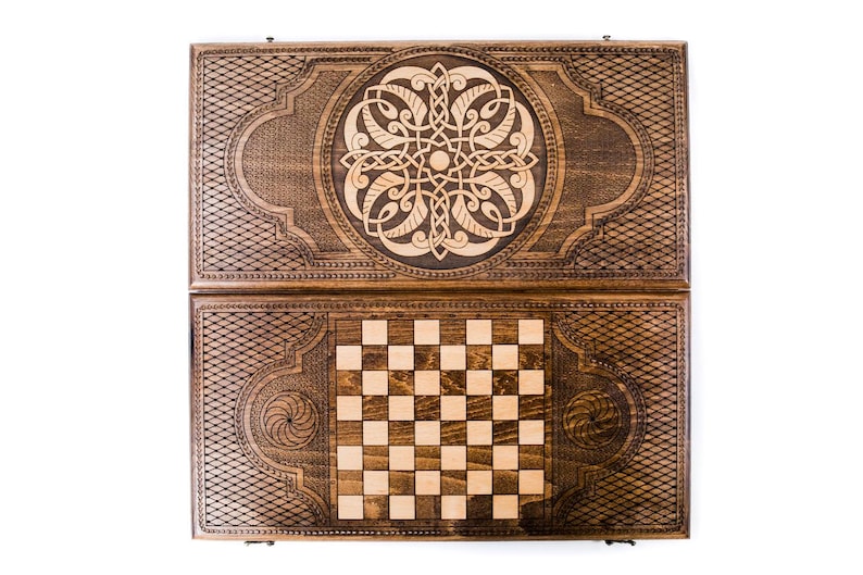 May include: A wooden backgammon and chess set. The dark brown wooden case is intricately carved with geometric and floral designs. The backgammon board is open, revealing a checkerboard pattern and a detailed central design.