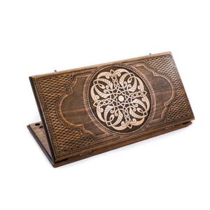 May include: A wooden backgammon board with intricate carved details. The board is a rich brown color, featuring a central circular design with a lighter wood inlay. The edges have a crosshatch pattern, and the board is open, ready for play.