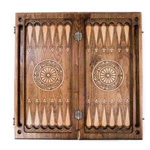 May include: A closed wooden backgammon board with detailed carved patterns. The board is split into two halves, each with a central circular design and alternating light and dark wood triangles. The board is ready for a game.