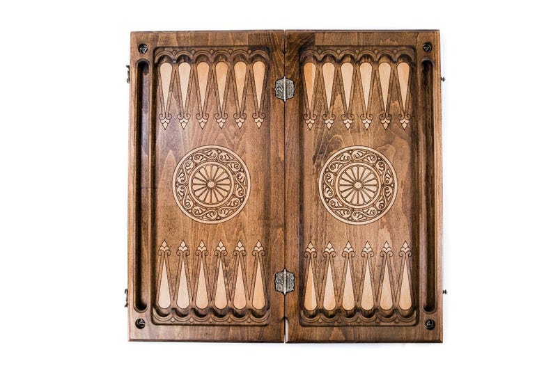 May include: A closed wooden backgammon board with detailed carved patterns. The board is split into two halves, each with a central circular design and alternating light and dark wood triangles. The board is ready for a game.