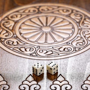 May include: A wooden backgammon board with a detailed circular design and two dice. The board is light brown with dark brown etched patterns. The dice are white with black dots.