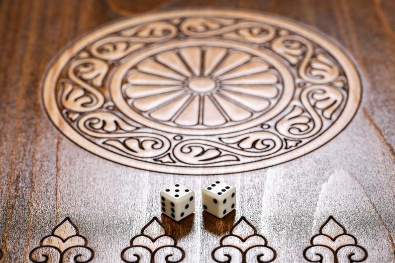 May include: A wooden backgammon board with a detailed circular design and two dice. The board is light brown with dark brown etched patterns. The dice are white with black dots.