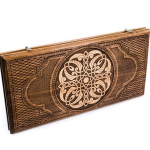 May include: A closed wooden backgammon board featuring a detailed Celtic knot design. The board is a warm brown colour with a lighter wood carving in the centre. The edges have a diamond pattern. Ready for play.