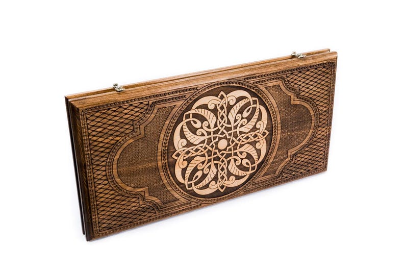 May include: A closed wooden backgammon board featuring a detailed Celtic knot design. The board is a warm brown colour with a lighter wood carving in the centre. The edges have a diamond pattern. Ready for play.