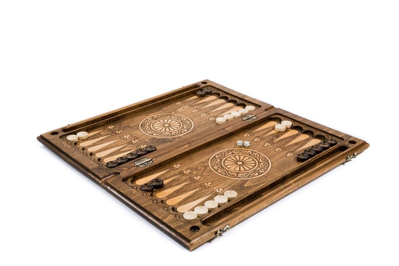 May include: A wooden backgammon board with a detailed, carved design. The board is brown with light and dark wood accents, and includes white and black game pieces, and dice. The game board is open and ready for play.