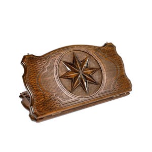 May include: A wooden backgammon board with a carved eight-pointed star in the center. The board is brown with intricate detailing and a textured surface. The edges are curved and the board is slightly open.