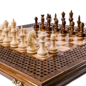 May include: A wooden chess set with light and dark pieces arranged on a checkered board. The board is set within a decorative, dark-stained wooden frame with a patterned border. The chess pieces are in their starting positions.