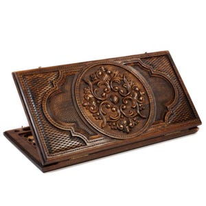 May include: A dark brown, wooden backgammon set with intricate carved floral designs. The set is open, revealing the playing surface with a diamond-patterned grid. The central design is a detailed, circular floral carving.