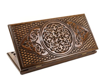 Puede incluir: Un tablero de backgammon rectangular de madera con intrincados patrones tallados a mano. La superficie de madera marrón oscuro muestra un motivo floral circular central flanqueado por volutas simétricas y una textura de rejilla de diamantes.