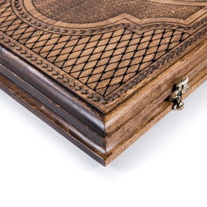 May include: Close-up of a wooden backgammon board. The board is made of light and dark brown wood with intricate carved details. The edges are dark brown, and the surface features a diamond pattern and decorative borders.