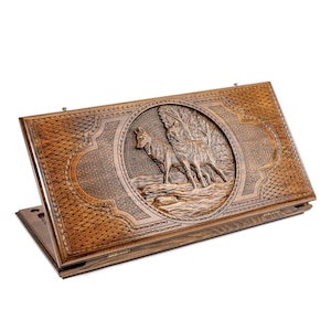 Puede incluir: Una caja de juego de madera con un diseño tallado de dos lobos en un entorno de bosque. La caja está cerrada y tiene una tapa con bisagras.