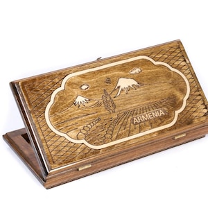 May include: Wooden box with a hinged lid, featuring a detailed engraved design. The design includes mountains, fields, and the word "ARMENIA" within a decorative border. The box is made of light brown wood.