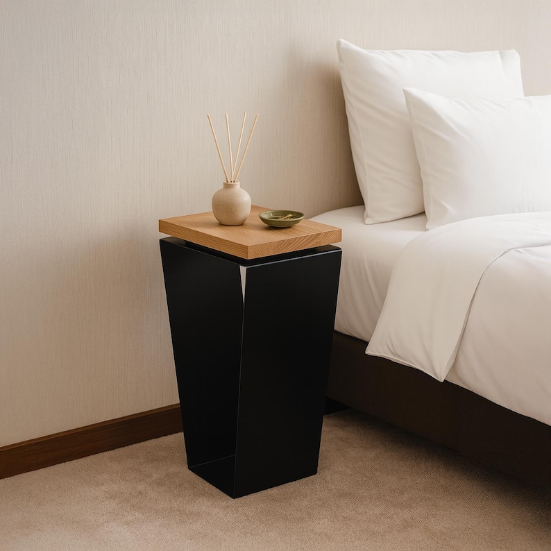 May include: A modern black side table with a natural wood top. The table has a geometric design and is next to a bed with white bedding. A small vase with reeds and a green dish sit on the table.