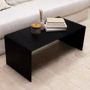 May include: A modern black coffee table with a minimalist design. The table has a rectangular top and curved legs. On the table are books, a candle in a glass jar with a wooden lid, and a marble diffuser with reeds. The table sits on a cream-colored rug.