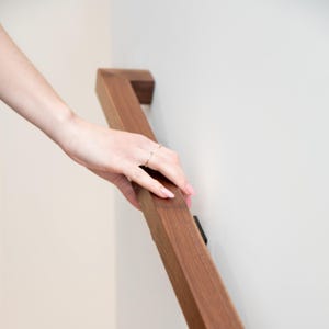 May include: A square-profile wooden handrail with a rich brown finish mounted to a plain white wall. The handrail features a smooth, straight design that runs diagonally across the frame, providing a sturdy grip for support along a staircase.