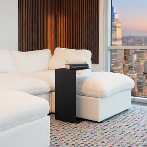 May include: A modern living room scene featuring a white sectional sofa, a black side table, and a patterned rug. Two books are on the side table. The background includes a wooden wall panel and a cityscape view through a large window.