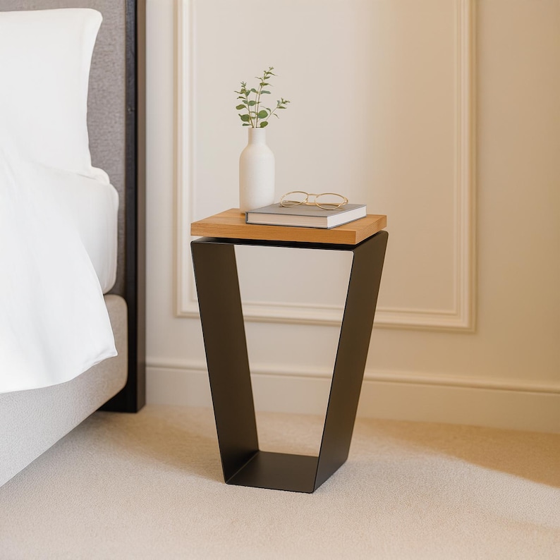 May include: A modern side table with a black metal base and a natural wood top. A white vase with greenery, a book, and glasses rest on the table. The table is next to a bed with white linens.