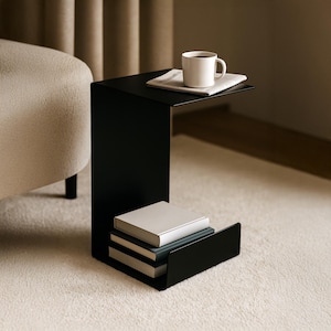 May include: A modern black side table with a C-shaped design. The table has a top surface holding a white mug of coffee and a newspaper. The lower section features a built-in shelf with a stack of books. The table is on a light-colored rug.