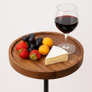 May include: A wooden serving tray with a selection of food and a glass of red wine. The tray holds strawberries, grapes, apricots, and a wedge of cheese. The wine glass is clear, and the tray is supported by a black metal stand.