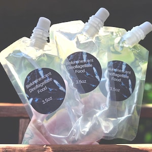 May include: Three clear plastic pouches with white spouts, each labeled "Bioluminescent Dinoflagellate Food" with a 3.5oz measurement. The labels are dark blue with a white and blue design. The pouches are partially transparent.