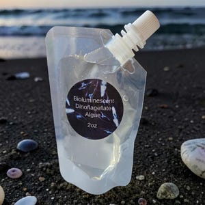 May include: A clear plastic pouch with a white screw cap containing Bioluminescent Dinoflagellate Algae. A circular black label on the front features the text Bioluminescent Dinoflagellate Algae 2oz in white lettering with small blue light streaks.