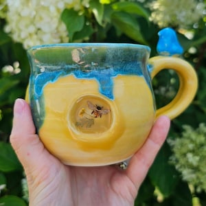 Pode incluir: Caneca de cerâmica artesanal com base amarela, borda azul e verde e alça amarela. A caneca apresenta um desenho de abelha e flor e um pequeno pássaro azul na alça. A caneca é segurada em uma mão.