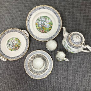 Peut inclure: Service à thé vintage avec un motif de paysage. L'ensemble comprend une théière, des assiettes, des bols et des tasses. Les bords sont ornés d'un motif gris et doré. Le centre de chaque pièce présente une scène de paysage colorée.