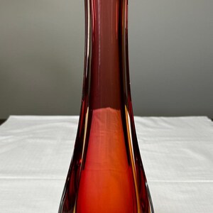 May include: A striking glass vase with a colour gradient. The vase shifts from a deep red at the top to orange and yellow at the base. Its elongated form and angled top give it a modern aesthetic. The vase is likely around 30 cm tall.