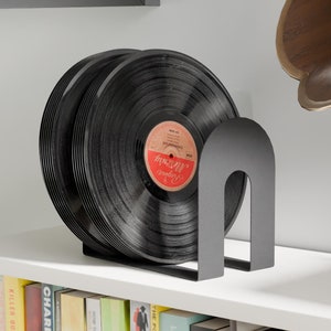 May include: A black metal record stand with a curved design holds a stack of black vinyl records. The stand is on a white shelf with a row of books below it.
