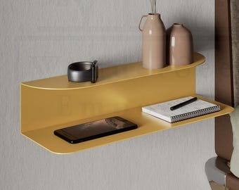 May include: A mustard yellow floating shelf with a curved design, mounted on a gray wall. The shelf holds a black watch, a smartphone, a notebook with a pen, and two beige vases with dried plants. A bed with a brown headboard is in the background.