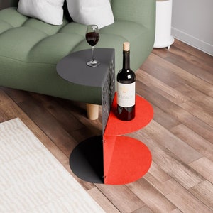 May include: A black and red metal side table with two tiers. The top tier has a glass of red wine and a bottle of red wine on it. The bottom tier is red. The table is in front of a green sofa.