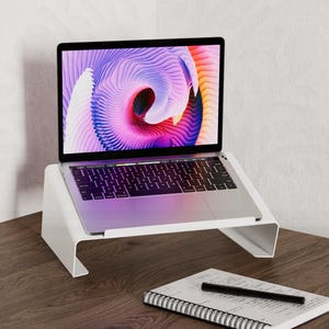 May include: A white laptop stand with a laptop on top of it. The laptop has a colorful abstract design on the screen. The stand is on a wooden desk with a notebook, a mouse, and a keyboard.
