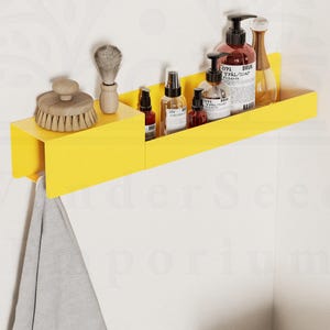 Puede incluir: Una estantería de metal amarilla con una toalla colgando debajo. La estantería está montada en una pared blanca y contiene varios artículos de tocador, incluyendo botellas de champú, acondicionador y gel de ducha, así como un cepillo y un peine.