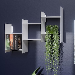 Puede incluir: Estantería de pared blanca y geométrica con libros y una planta en maceta. Los estantes están dispuestos en un diseño asimétrico, con libros a la izquierda y una planta verde colgante a la derecha. La pared es azul oscuro.