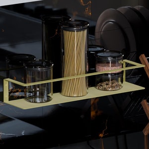 Może przedstawiać: Złota metalowa półka z czterema szklanymi słoikami wypełnionymi różnymi produktami, w tym makaronem, przyprawami i suszonymi produktami. Półka jest zamontowana na czarnej, wyłożonej kafelkami ścianie.