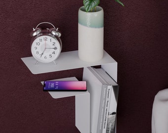 May include: White metal wall shelf with two tiers. The top tier has a small potted plant and a silver alarm clock. The bottom tier has a smartphone and a stack of books. The shelf is mounted on a burgundy wall.