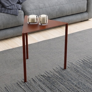 Puede incluir: Una escena de salón moderno con un sofá gris con cojines y una manta. Una pequeña mesa auxiliar triangular, de color marrón rojizo, sostiene dos velas. Una alfombra gris con un patrón lineal completa la decoración.