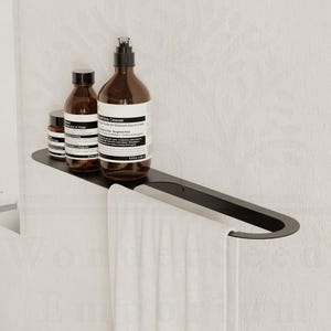 May include: A black metal shelf with a towel rail, holding three brown bottles. A white towel with a grey stripe hangs from the rail. The shelf is mounted on a white wall, creating a minimalist bathroom aesthetic.