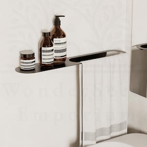May include: A modern black bathroom shelf holding brown skincare bottles. A white towel hangs below the shelf. The bathroom features a white sink and a mirror.