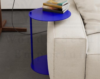 May include: A blue side table with a round top and a single leg. The table is next to a white couch with a stack of books on top.