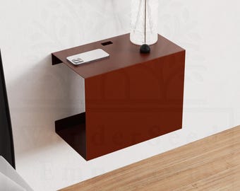 Puede incluir: Una mesita de noche moderna, montada en la pared, en un tono marrón rojizo oscuro. El diseño minimalista presenta una parte superior rectangular con una ranura y un estante inferior. Un smartphone y un jarrón con flores secas están encima. La mesita mide 30 cm de ancho.