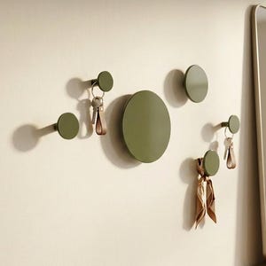May include: Olive green wall hooks in various sizes, some holding keys and a scarf. The hooks are mounted on a light-colored wall, with a full-length mirror and wooden cabinet in the background. A modern and minimalist design.