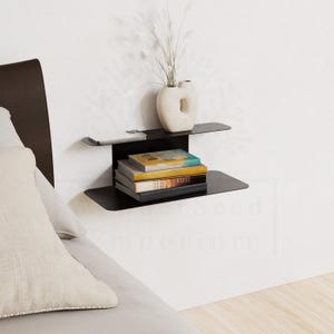 May include: A black metal floating shelf mounted on a white wall, holding books and a cream-colored vase with dried plants. The shelf has a minimalist design, with two tiers for storage. The books have colorful covers.