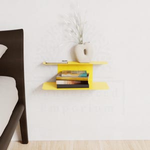 May include: A bright yellow floating shelf with two levels, mounted on a white wall. The top shelf holds a smartphone and a cream-colored vase with white decorative stems. The lower shelf displays a stack of books.