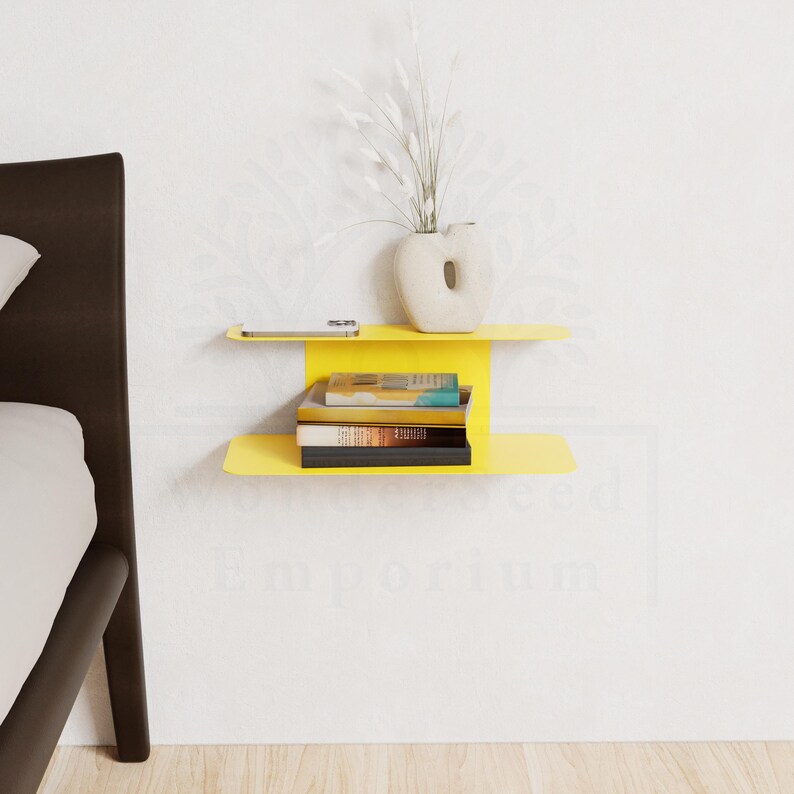 May include: A bright yellow floating shelf with two levels, mounted on a white wall. The top shelf holds a smartphone and a cream-colored vase with white decorative stems. The lower shelf displays a stack of books.