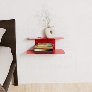 May include: A modern, red metal wall shelf with two tiers. The top shelf holds a small white vase with dried plants and a small white object. The bottom shelf displays a stack of books. The shelf is mounted on a white wall.