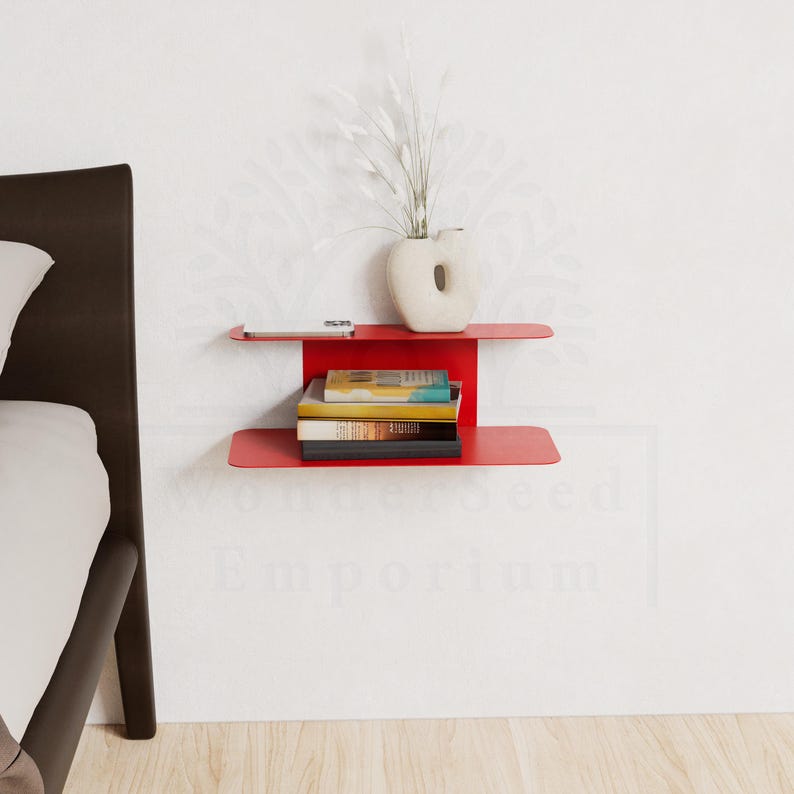 May include: A modern, red metal wall shelf with two tiers. The top shelf holds a small white vase with dried plants and a small white object. The bottom shelf displays a stack of books. The shelf is mounted on a white wall.